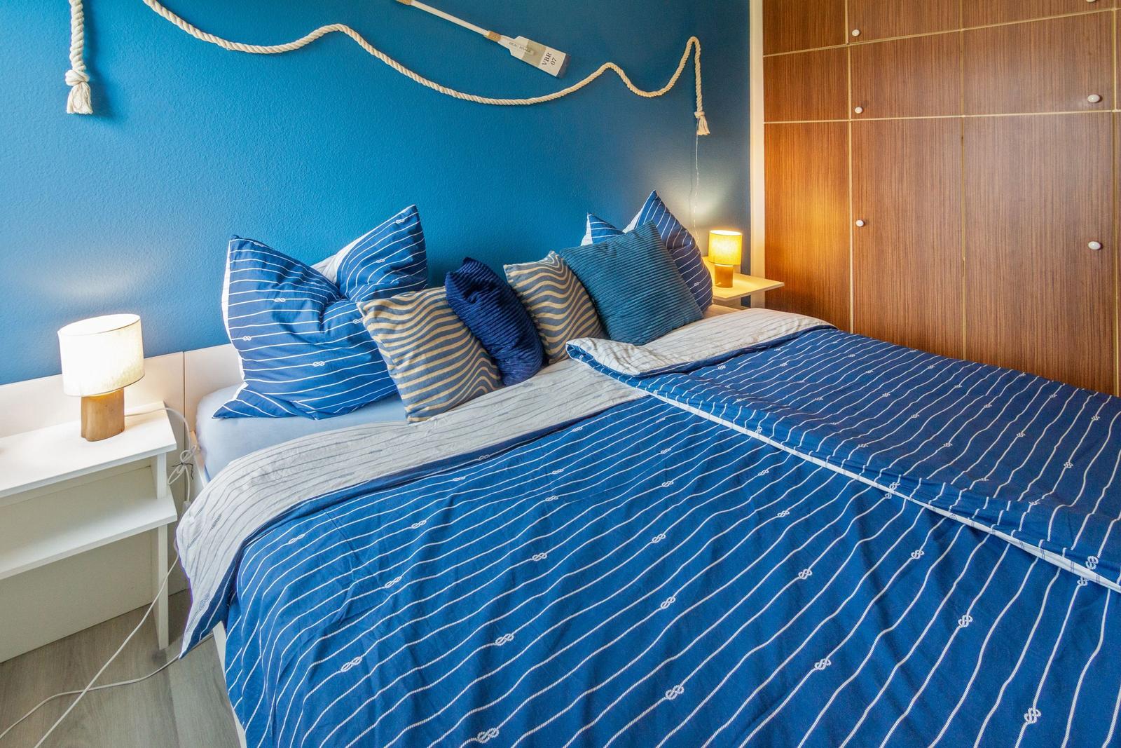 Schlafzimmer mit blauer Wand, Bett mit blau-weißem Bettzeug und Holzschrank.