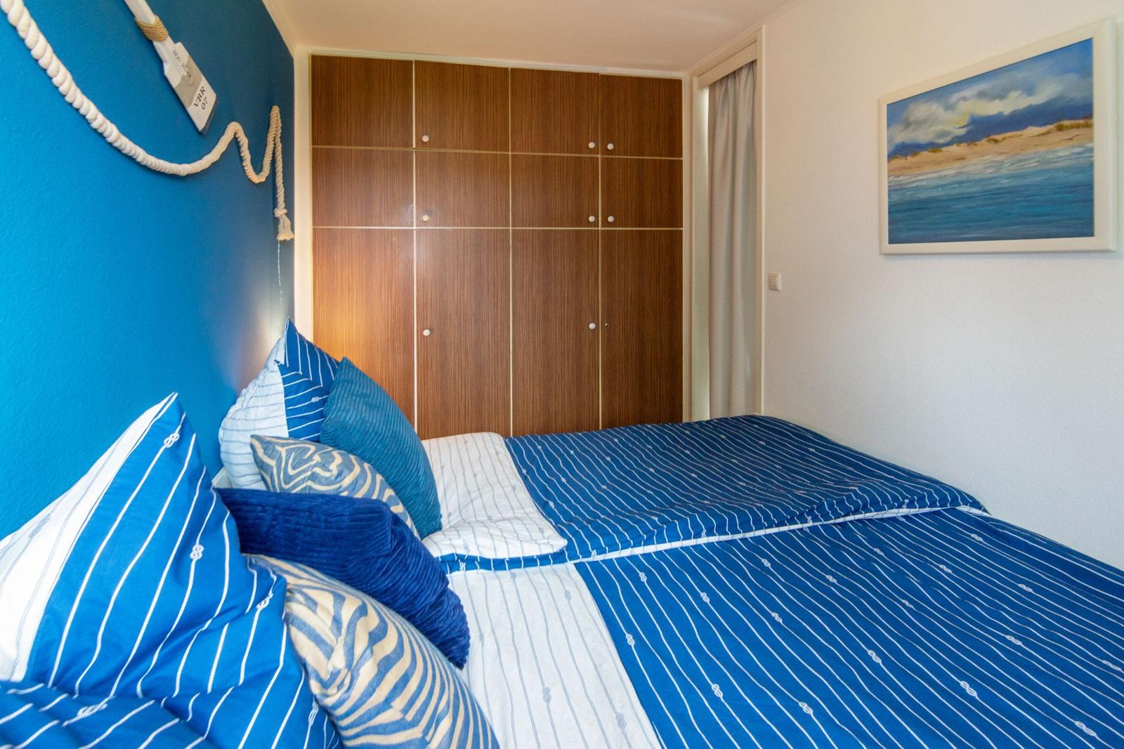 Schlafzimmer mit Bett, Holzschrank und Wandbild. Wand ist blau mit Seildekoration.