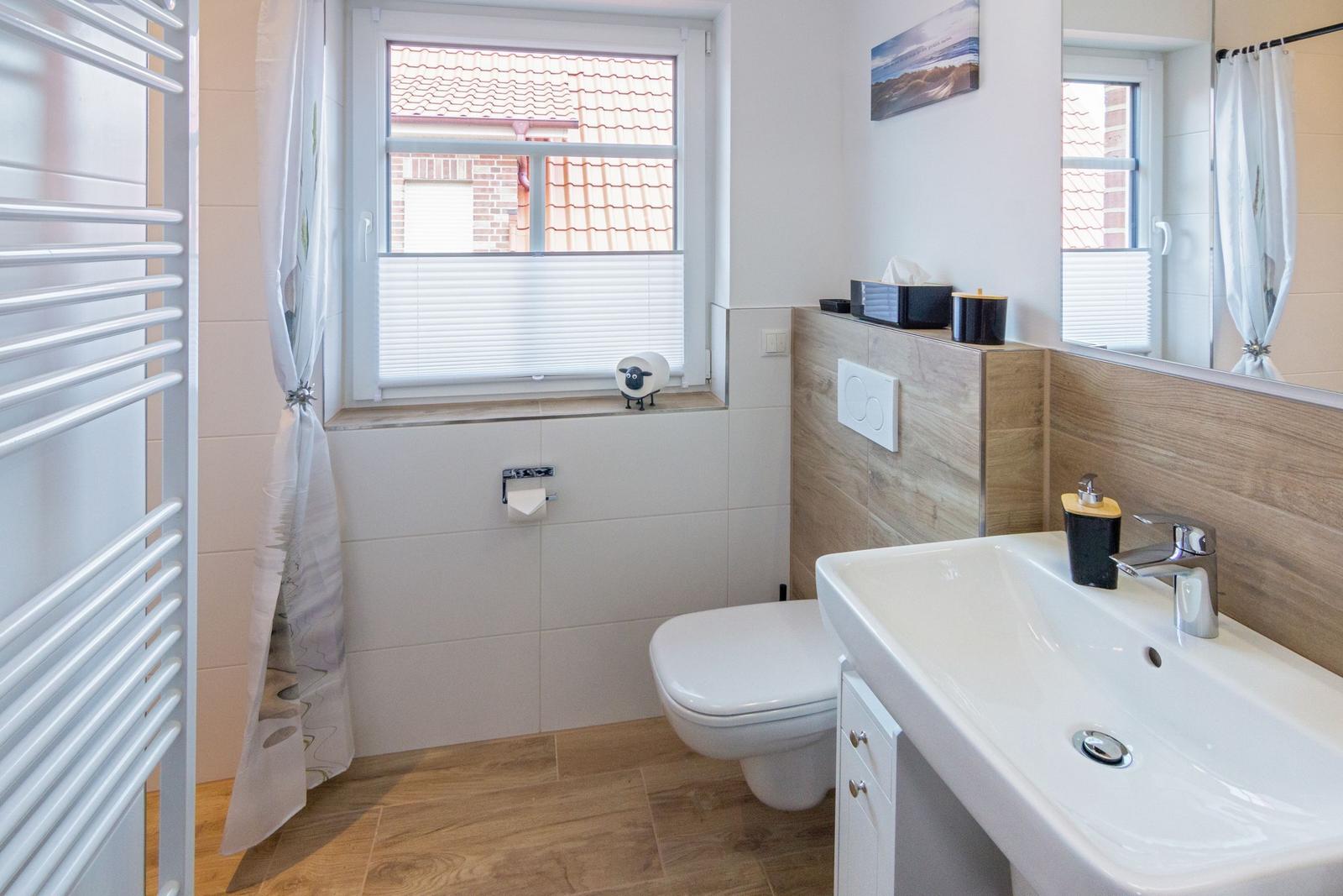 Badezimmer mit Toilette, Waschbecken und Fenster. Holzoptik und weißer Fliesenbelag.