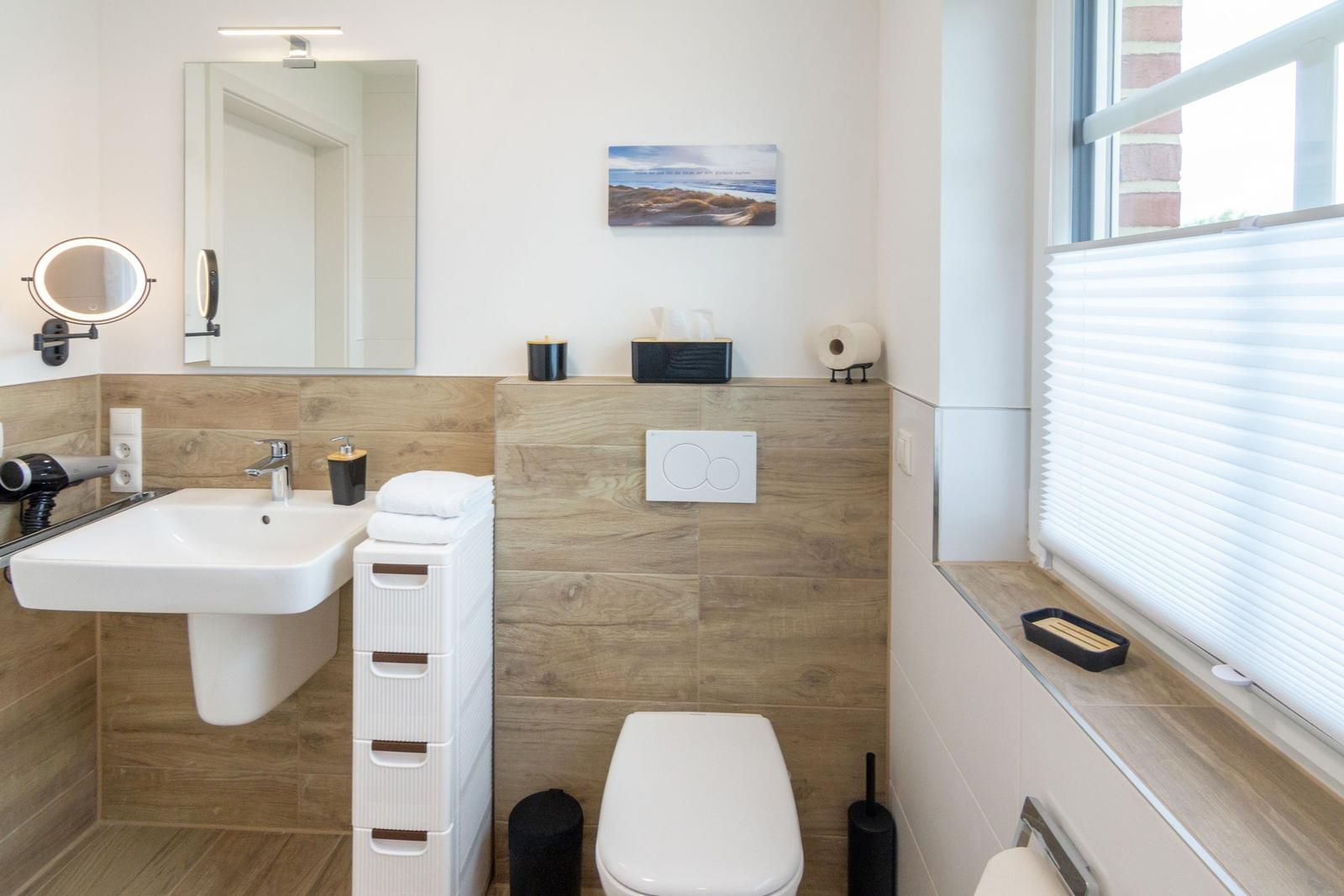 Badezimmer mit Waschbecken, Spiegel, Toilette und Fenster mit Jalousie.