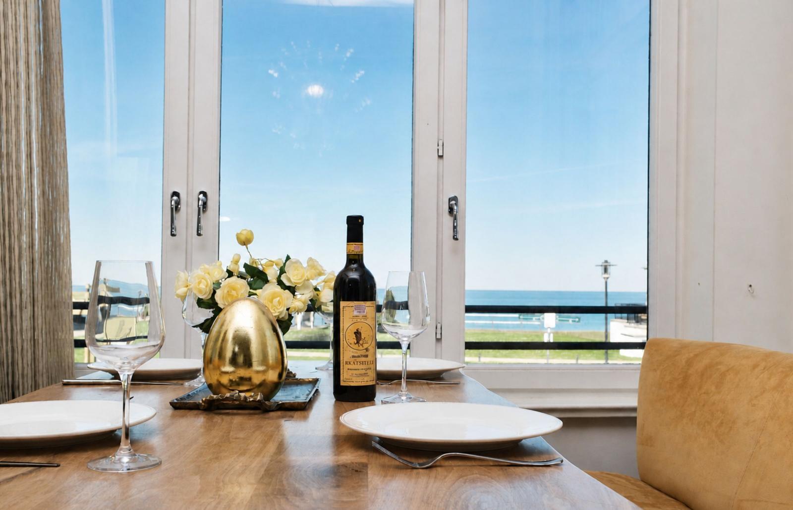 Esstisch mit Weinglas, Flasche und Blumen vor Fenster mit Meerblick.