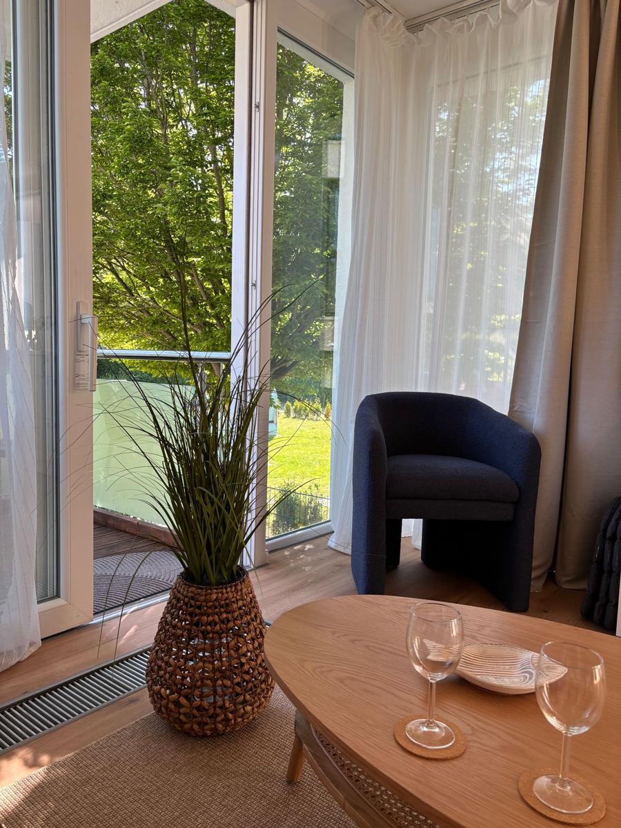 Ein Raum mit blauem Sessel, Holztisch und Pflanze vor großem Fenster mit Blick auf Grün.