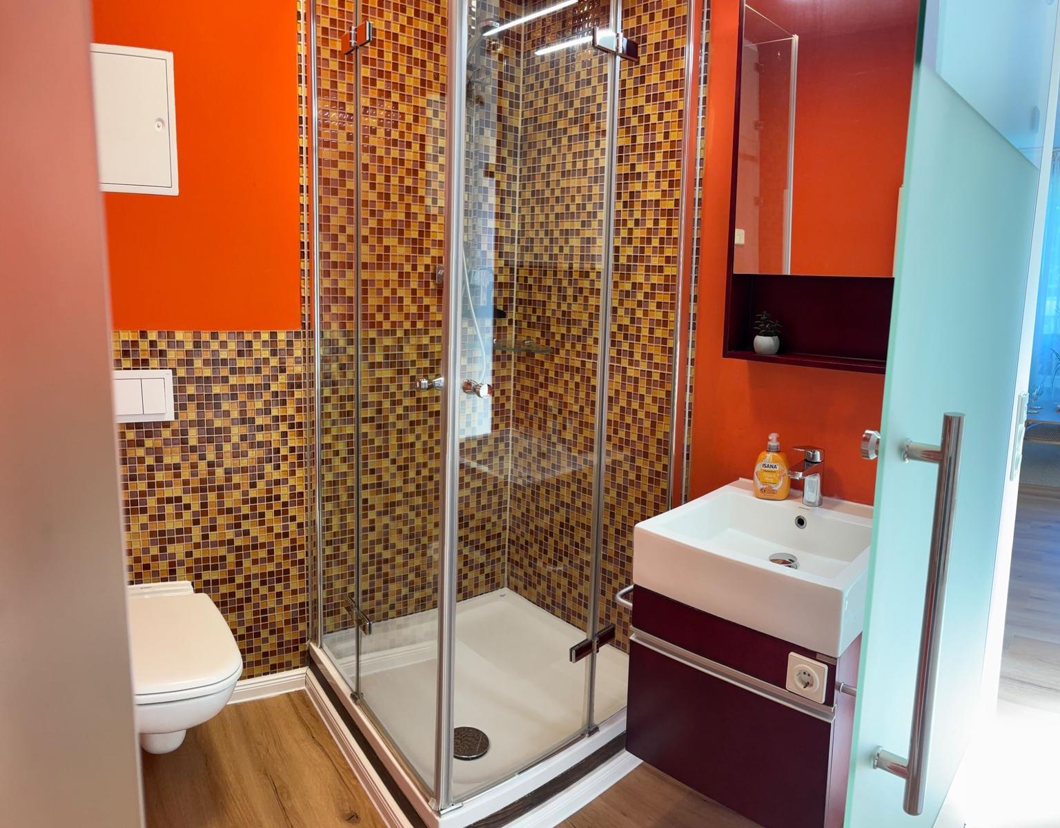 Badezimmer mit Dusche, Waschbecken und Toilette. Mosaikfliesen und orange Wand.