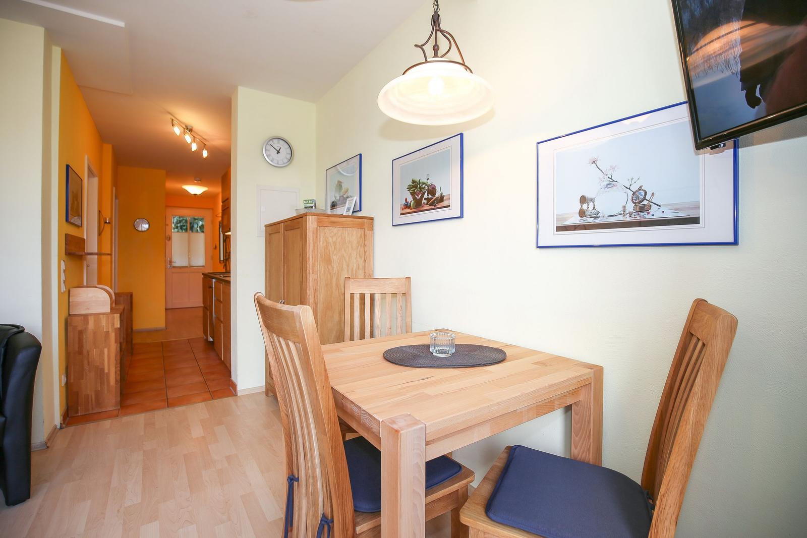 Essbereich mit Holztisch und Stühlen, Wandbilder und TV. Hintergrund: Küche und Flur.