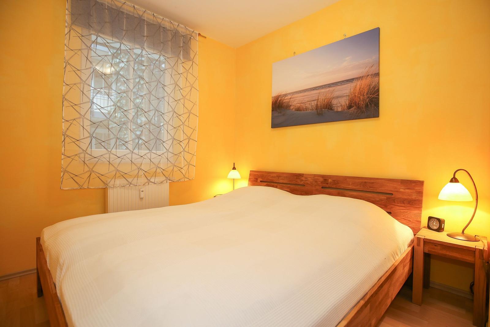 Schlafzimmer mit Holzbett, gelben Wänden, Fenster mit Vorhang und Wandbild.