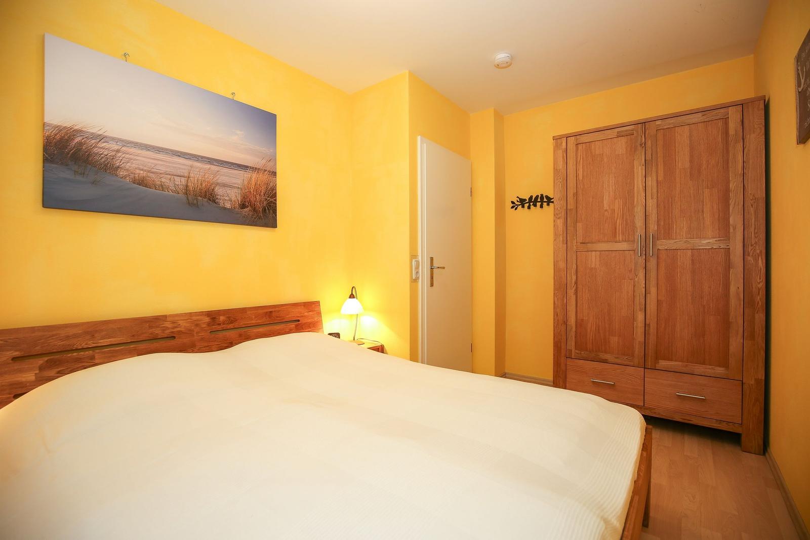 Schlafzimmer mit gelben Wänden, Holzbett und Kleiderschrank. Wandbild zeigt Strand.