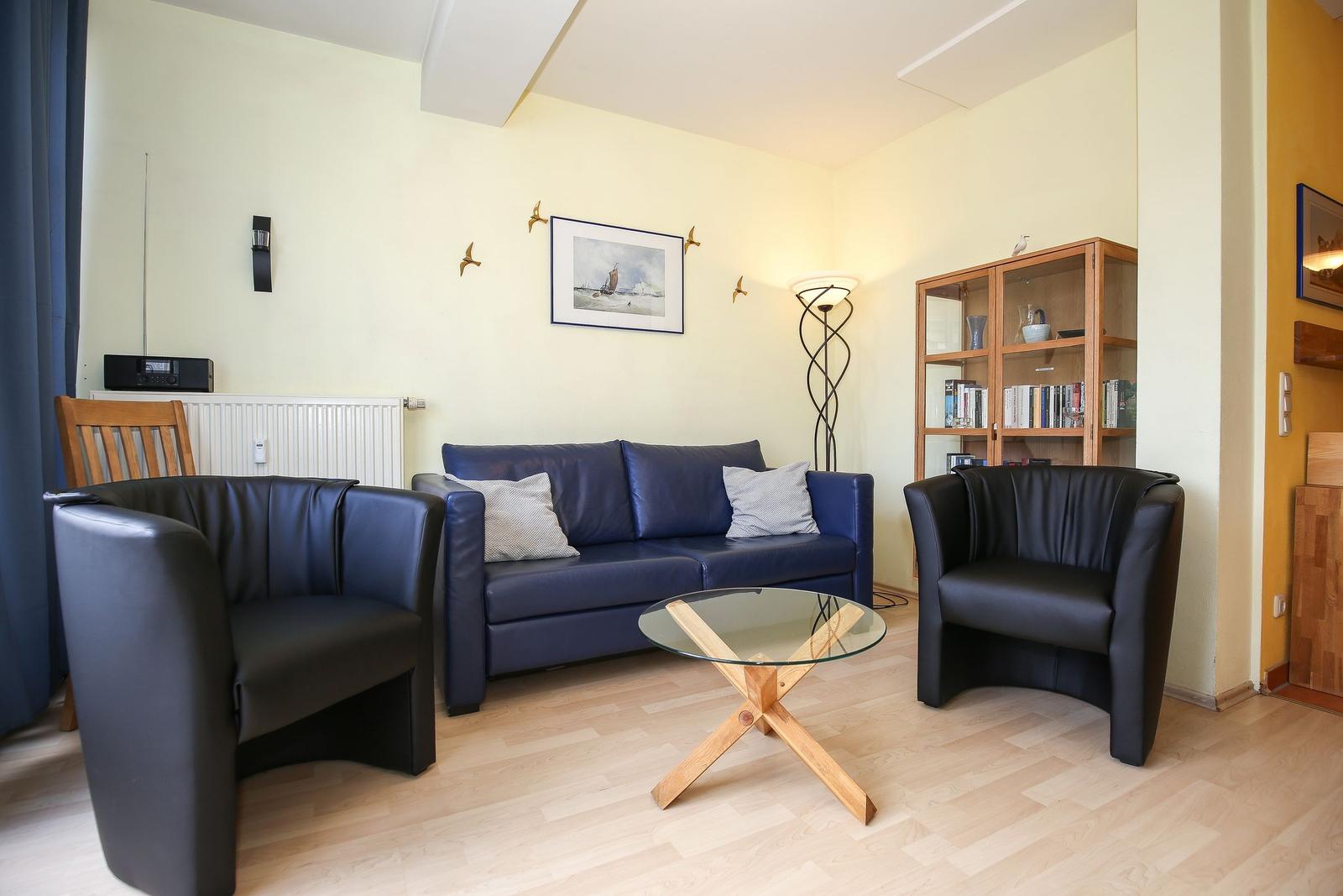 Ein Wohnzimmer mit blauem Sofa, zwei schwarzen Sesseln und einem Glas-Tisch.
