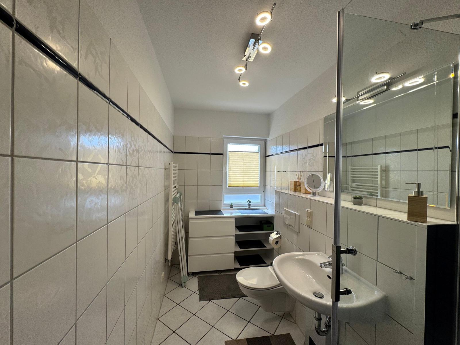 Badezimmer mit Dusche, Waschbecken, WC und Fenster.