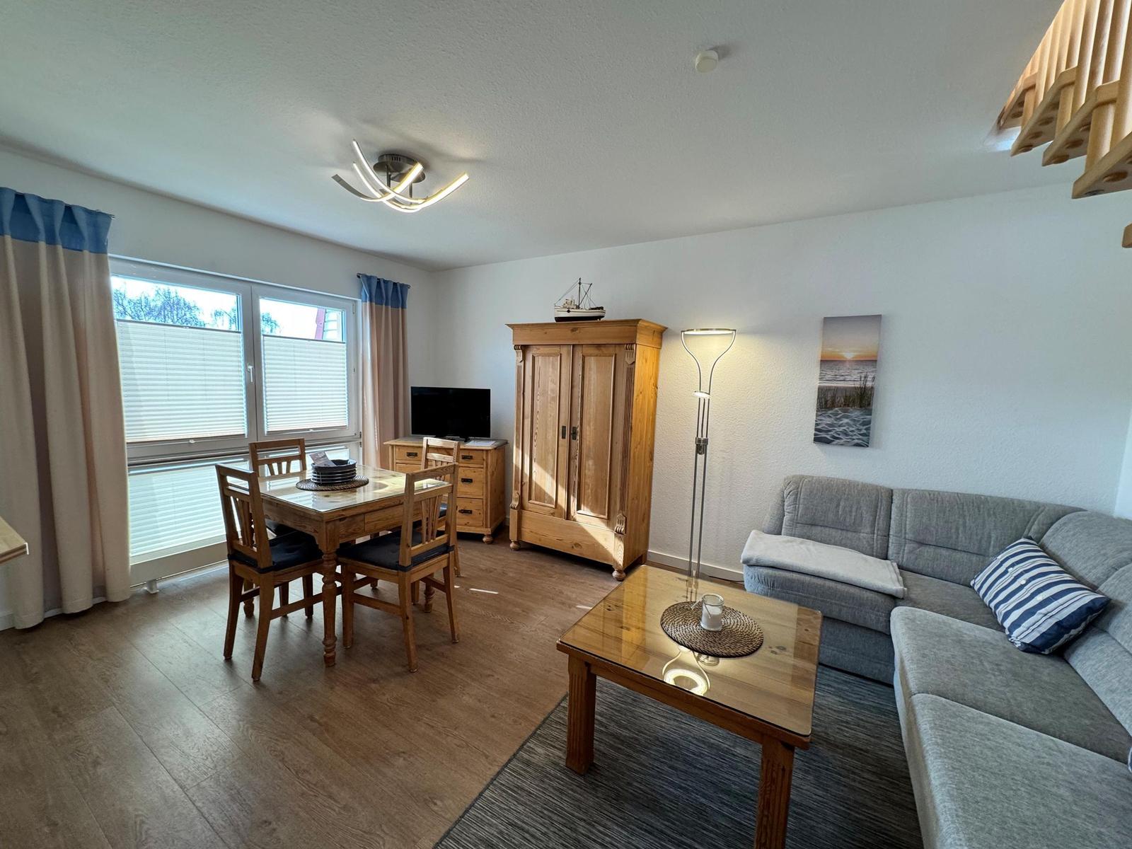 Wohn- und Essbereich mit Sofa, Tisch, Stühlen und Holzmöbeln. Fenster mit Jalousien und Vorhängen.