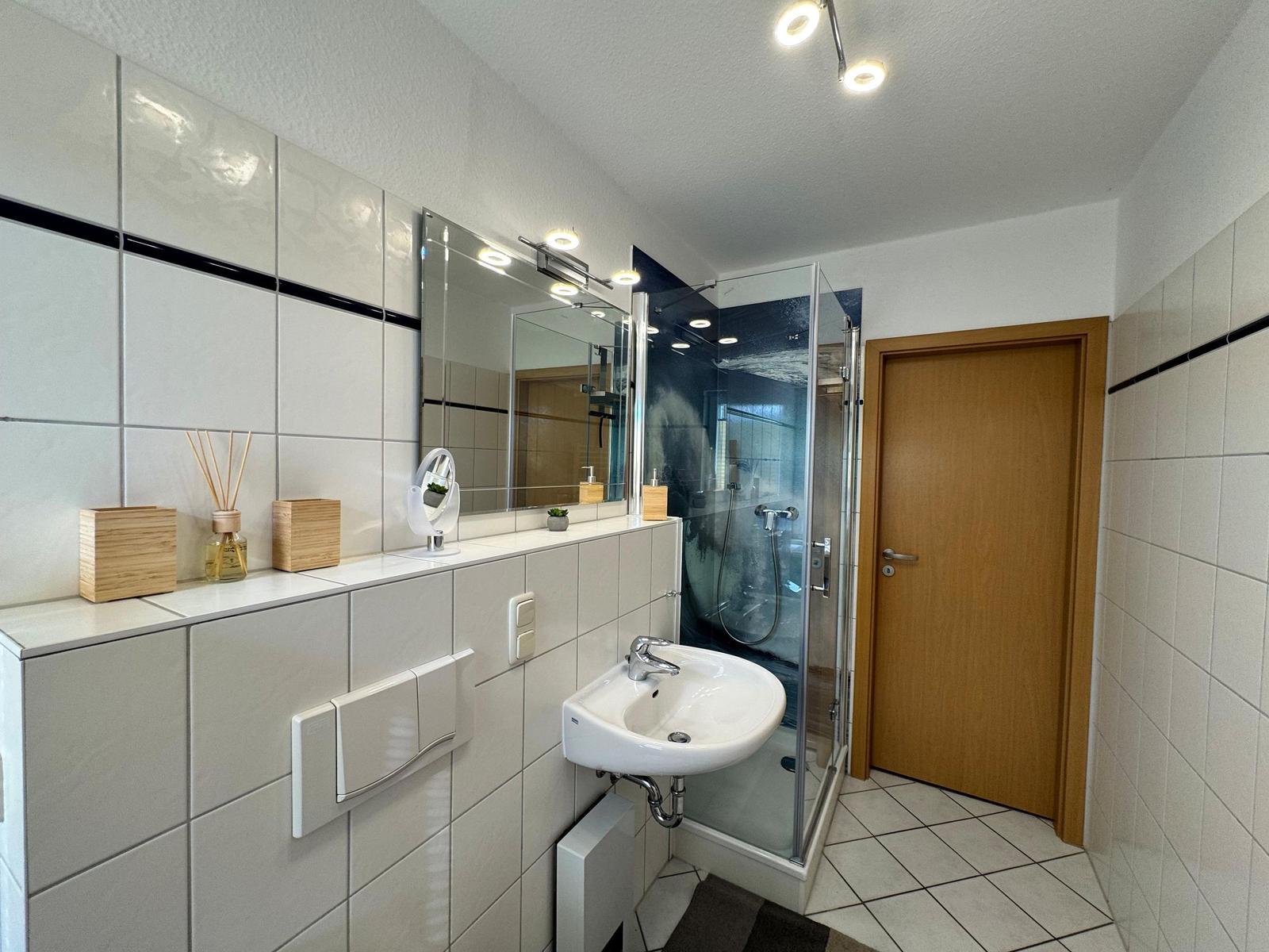 Badezimmer mit Dusche, Waschbecken und Spiegel. Fliesenwand und Holztür sichtbar.