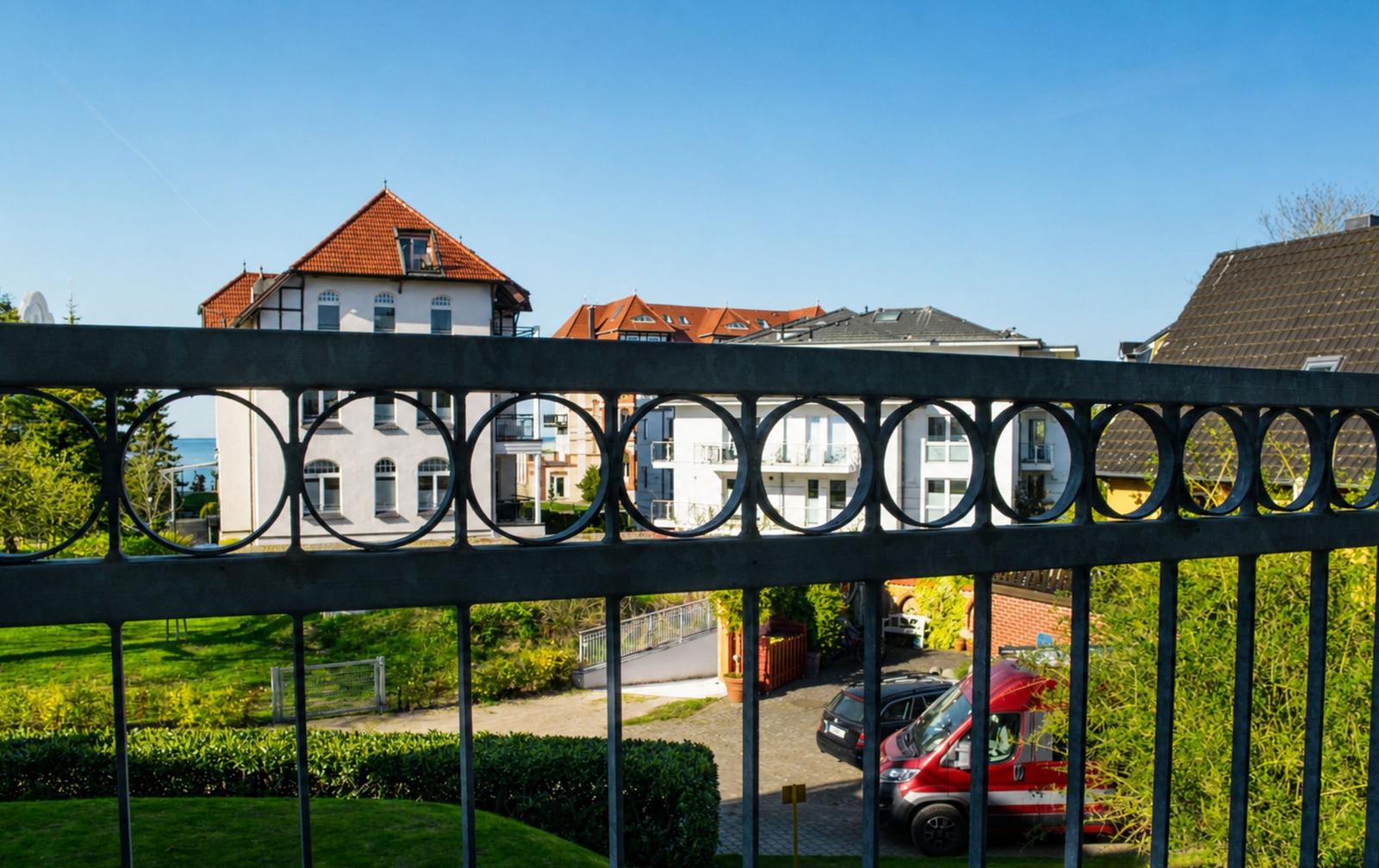 Durch einen Metallzaun blickt man auf Wohnhäuser und einen Garten mit einem roten Van.