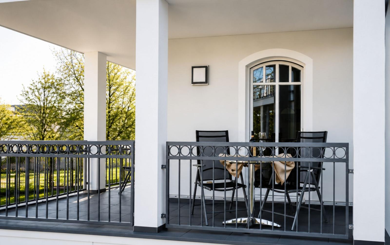 Terrasse mit Tisch, zwei Stühlen und Gittergeländer. Hintergrund: grüner Garten und Fenster.