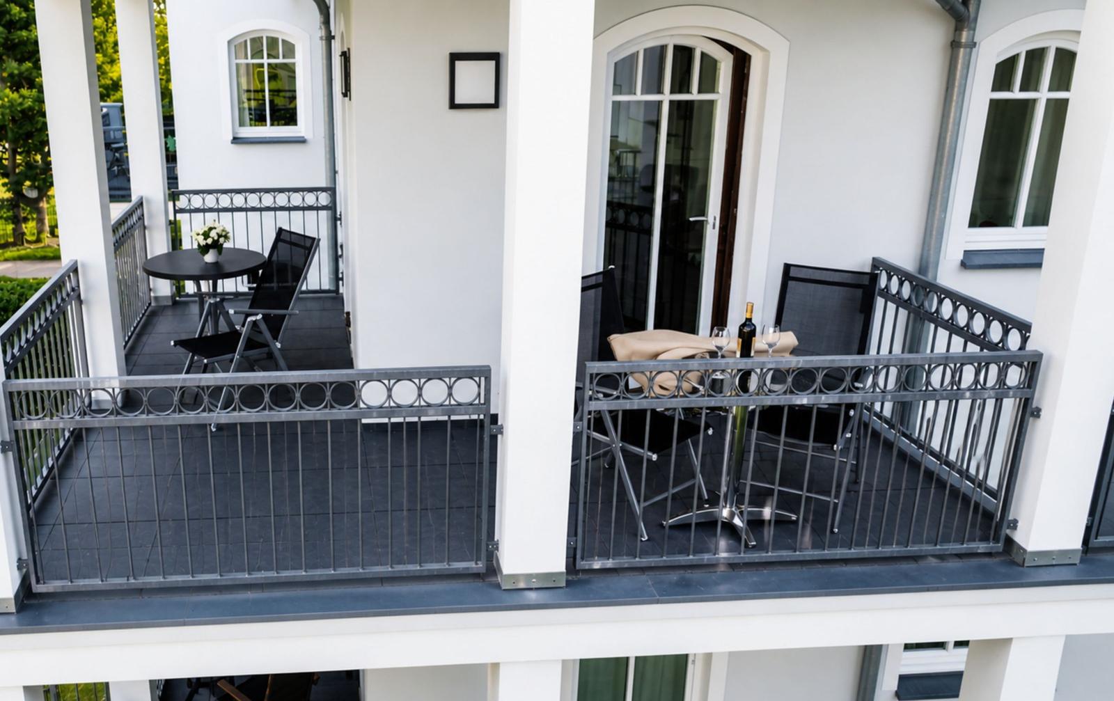 Terrasse mit Tisch, Stühlen und Weinflasche. Weiße Hauswand und schwarz geflieste Fläche.