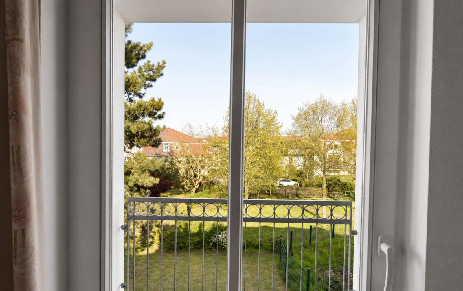Durch die offene Tür blickt man auf einen Balkon mit Geländer und grüne Bäume im Hintergrund.
