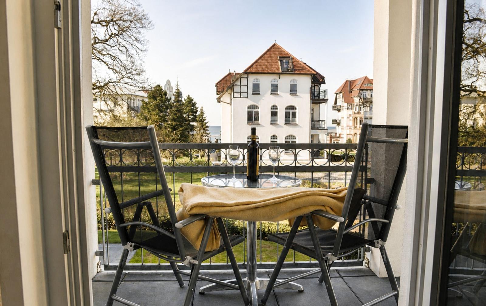 Terrasse mit Tisch, zwei Stühlen und Flasche Wein. Blick auf Gebäude und Grünfläche.