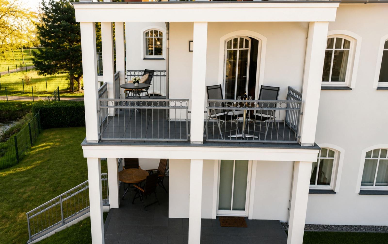 Terrasse mit Tischen und Stühlen, umgeben von grünem Garten und weißen Säulen.