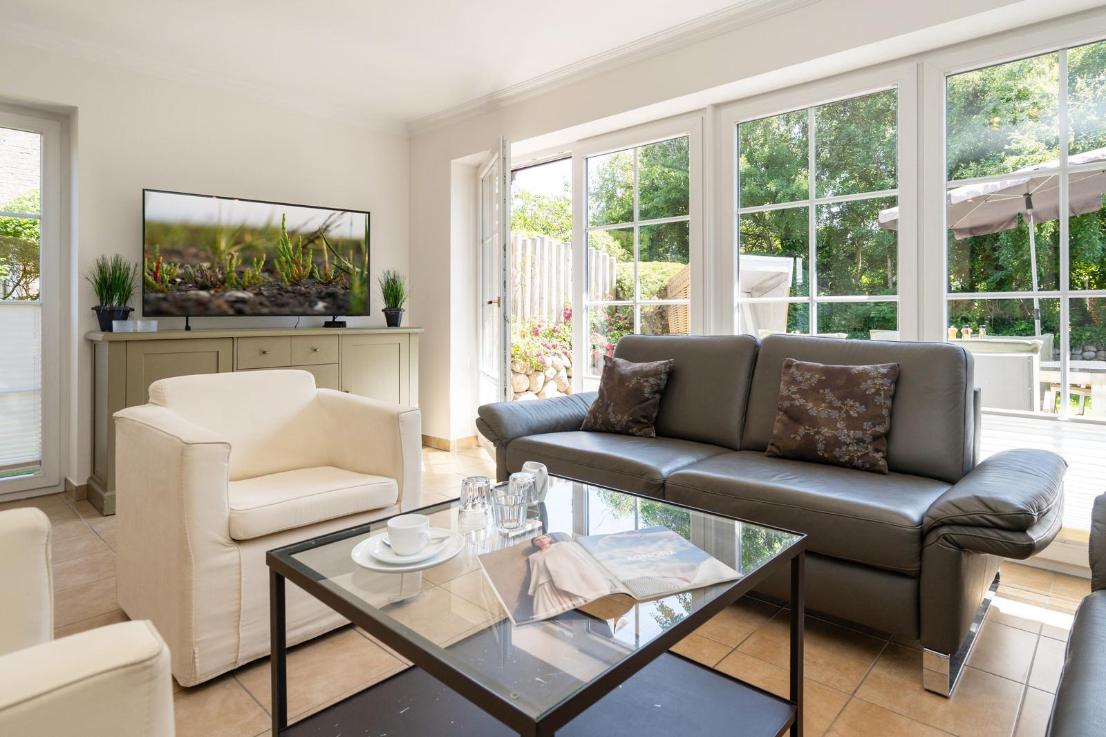 Wohnzimmer mit Sofa, Sessel und TV. Große Fenster mit Blick auf Garten.
