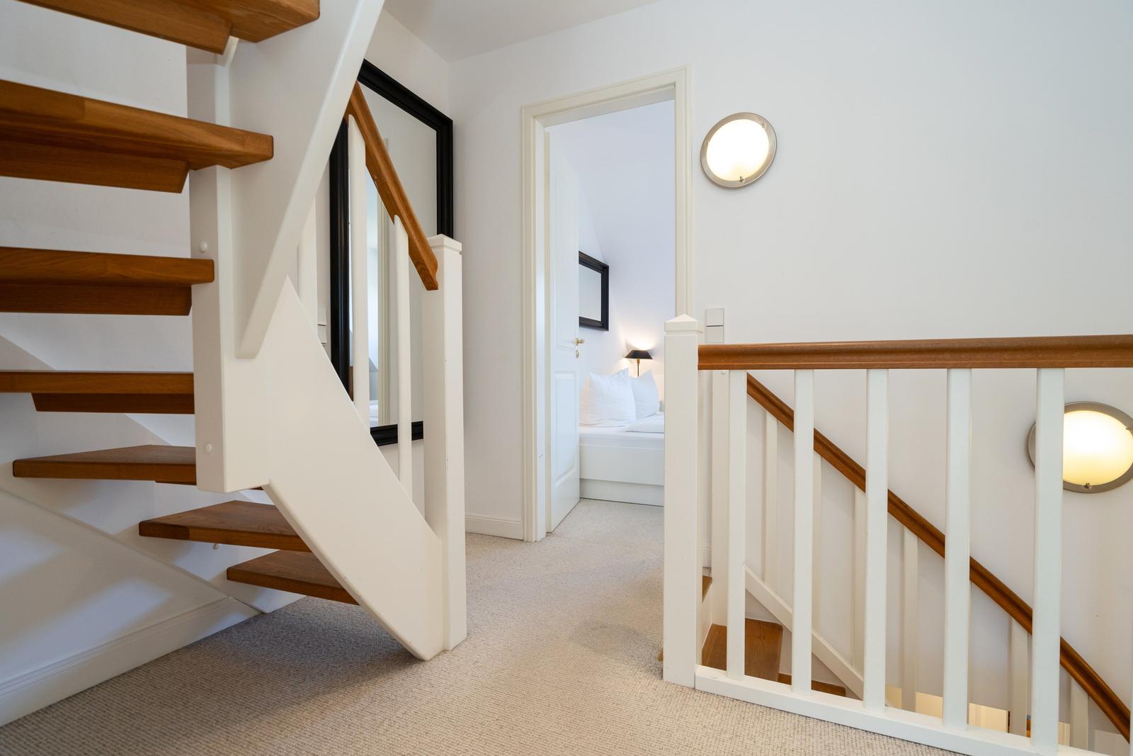 Treppenhaus mit Holzstufen und weißer Balustrade. Im Hintergrund ist ein Schlafzimmer mit Bett sichtbar.
