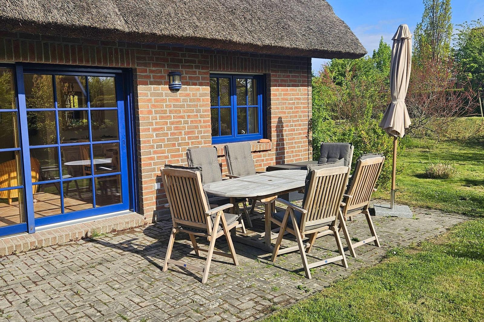 Terrasse mit Tisch und Stühlen vor einem Haus mit strohgedecktem Dach und blauen Fenstern.