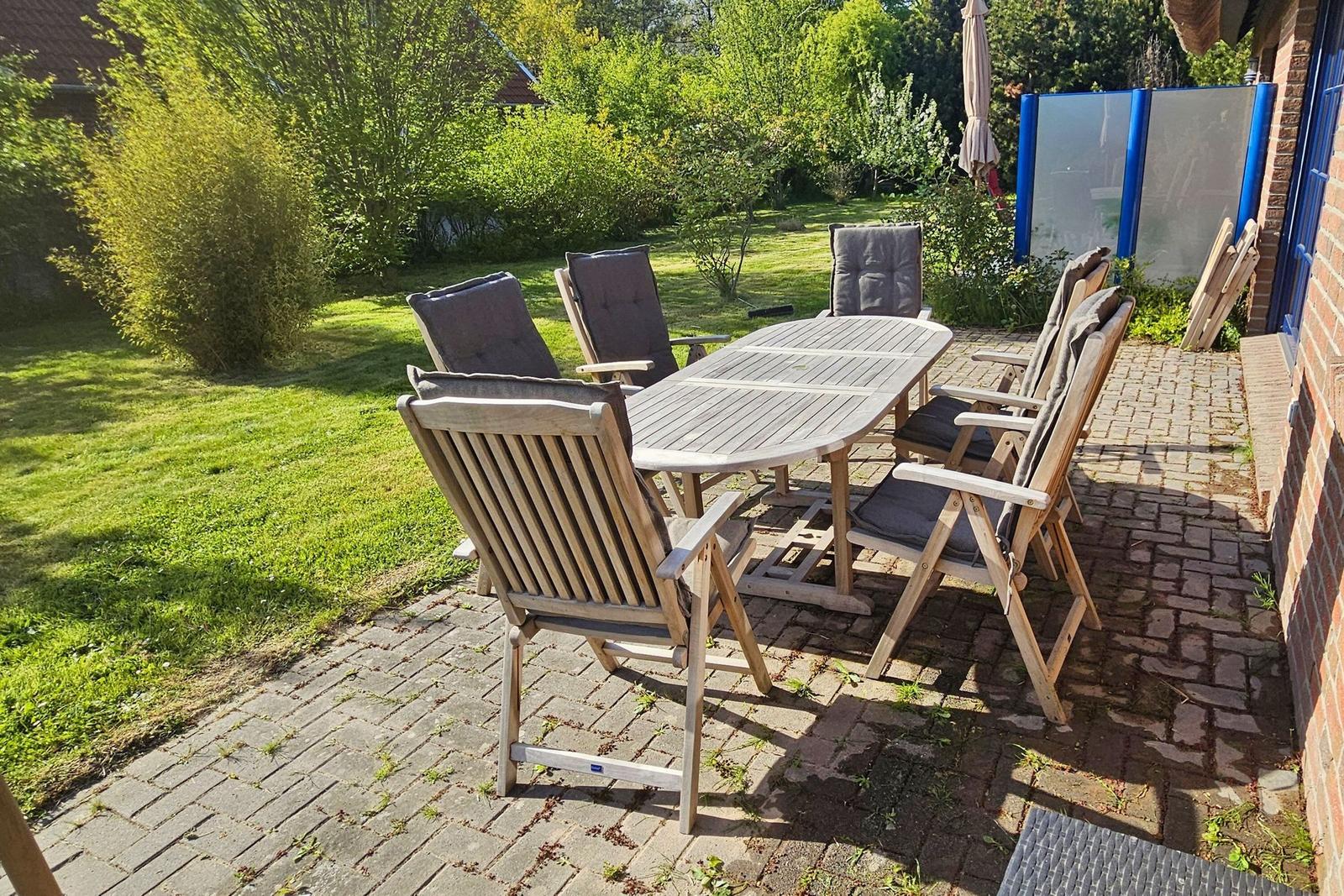 Holztisch mit sechs Stühlen auf Steinplatten. Hintergrund: Gras und Bäume.