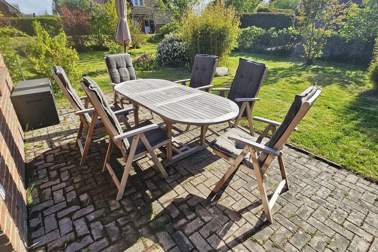 Terrasse mit Holztisch und sechs Stühlen mit Kissen, umgeben von Grünflächen.