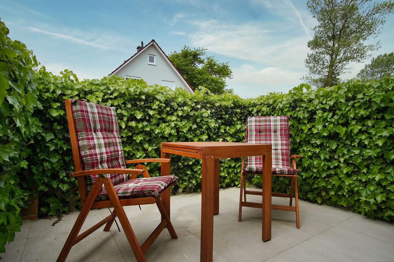 Hinter einem grünen Heckenzaun steht ein Holztisch mit zwei Stühlen mit karierten Sitzkissen.