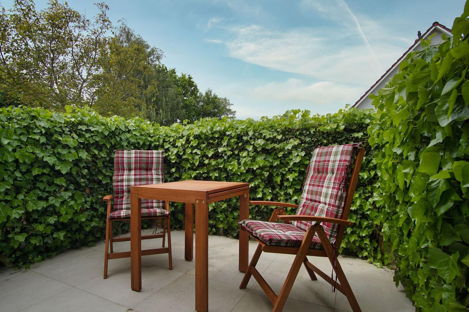Terrasse mit Holztisch und zwei Stühlen mit karierten Sitzkissen, umgeben von grünen Sträuchern.