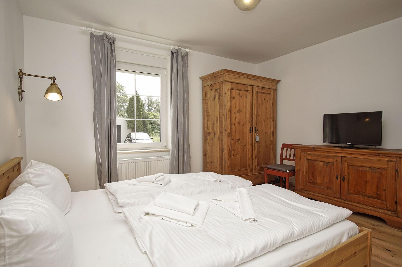 Schlafzimmer mit Bett, Kleiderschrank und TV. Fenster mit Vorhängen. Holzboden und Wandlampen.