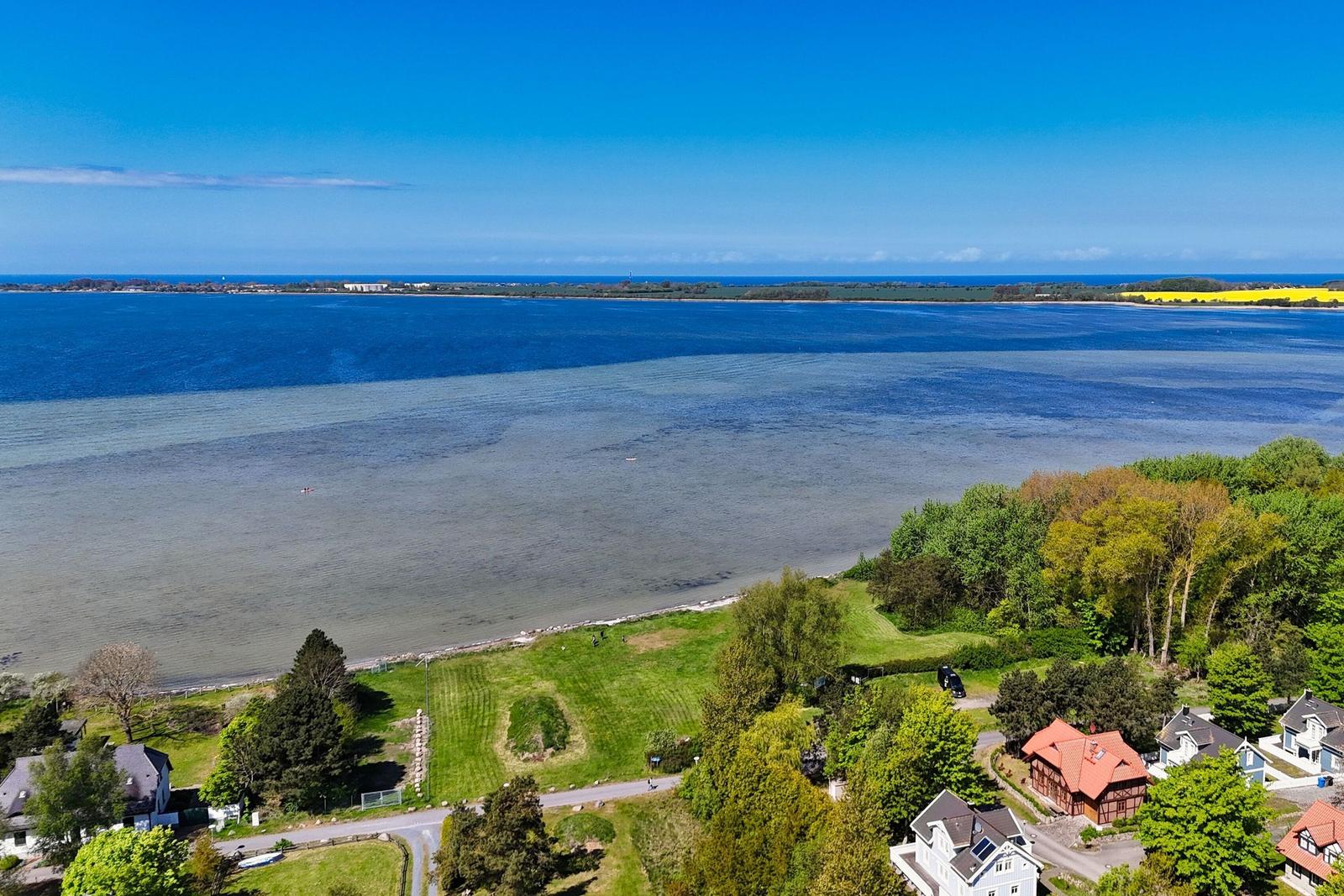 Ausblick auf einen See mit Uferbereich, Grünflächen und Häusern unter blauem Himmel.