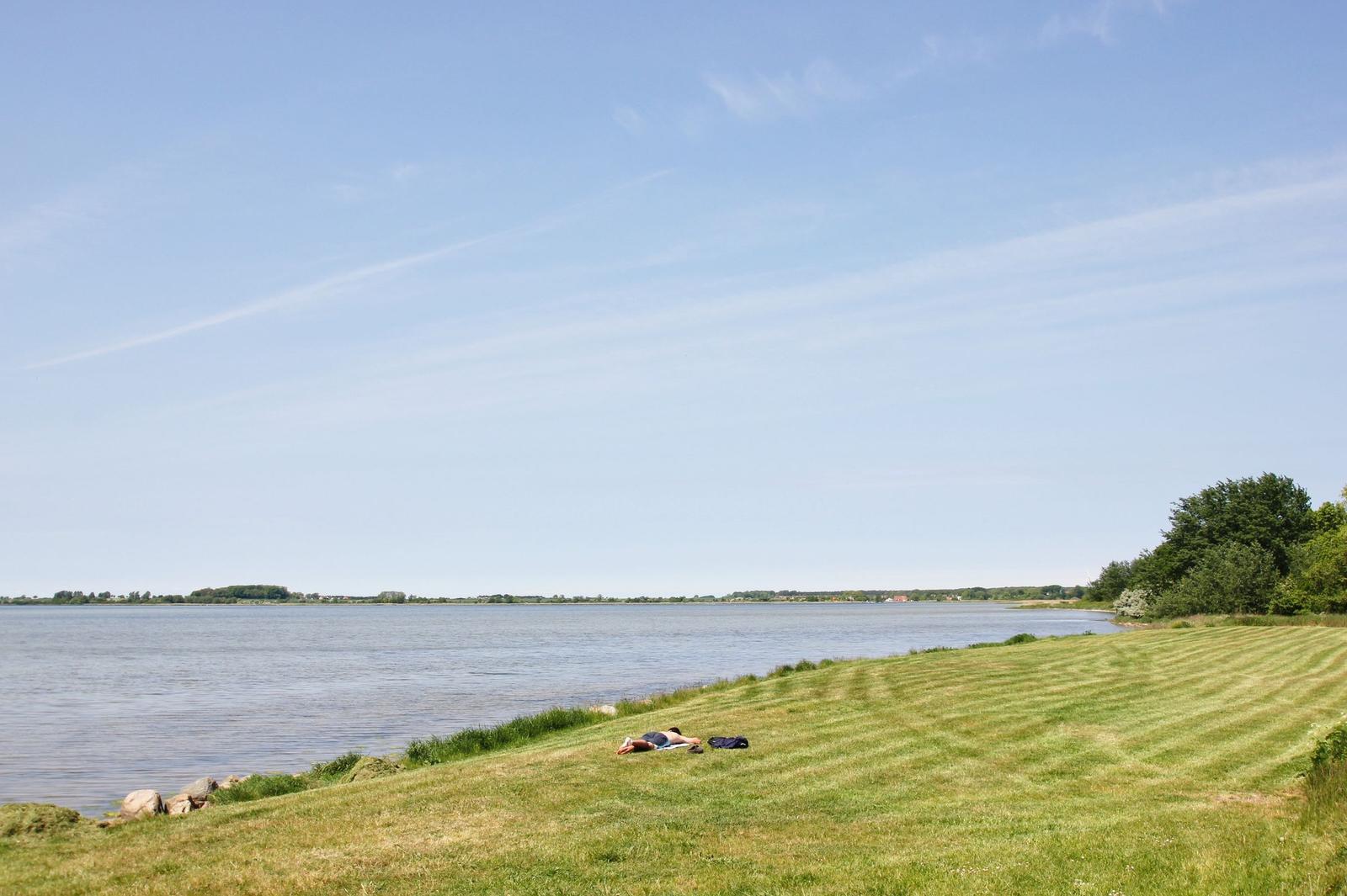 Ein Mensch liegt auf einer grünen Wiese am Ufer eines Sees unter blauem Himmel.