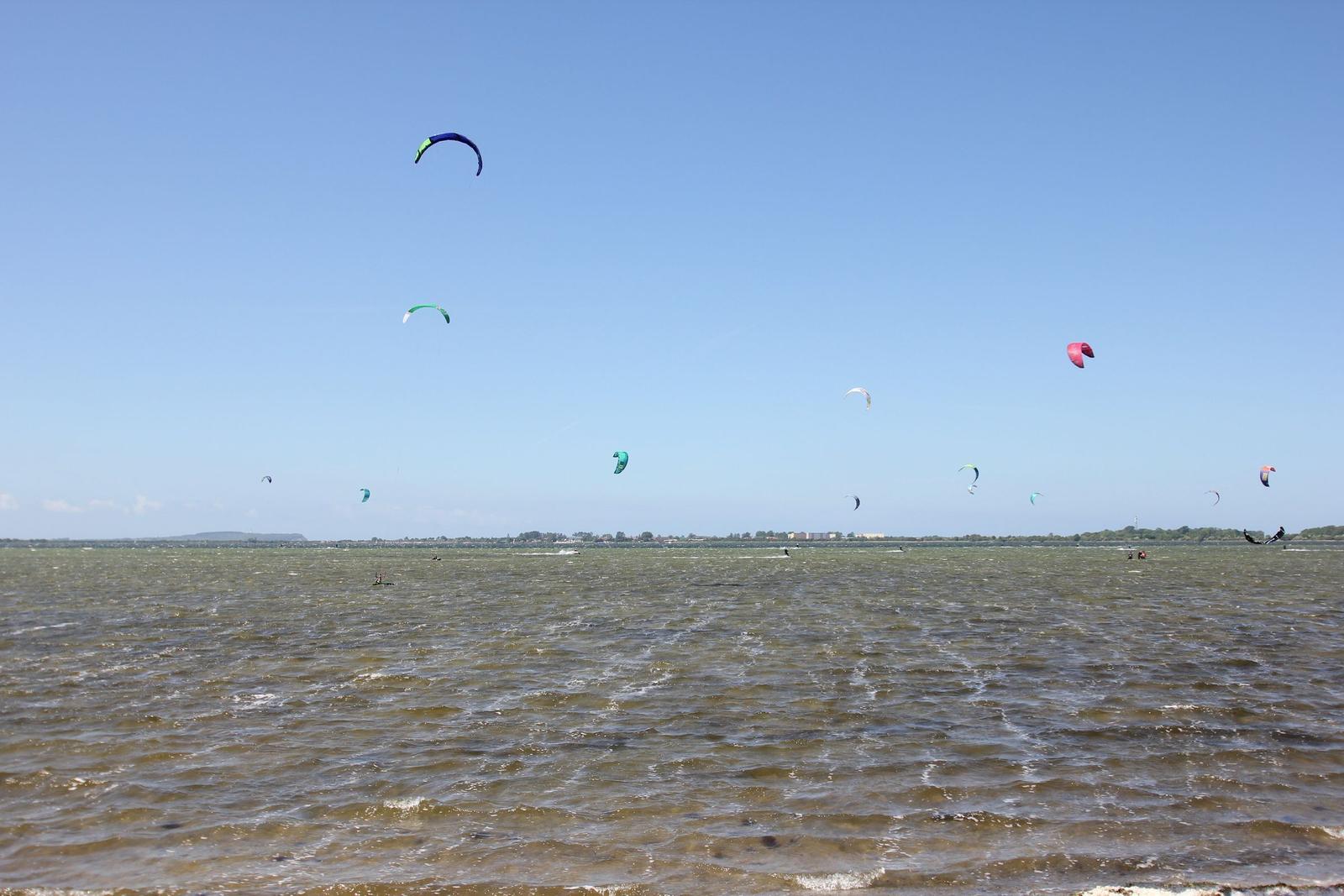 Mehrere Kitesurf-Paraschen fliegen über das Wasser. Menschen surfen auf dem See.