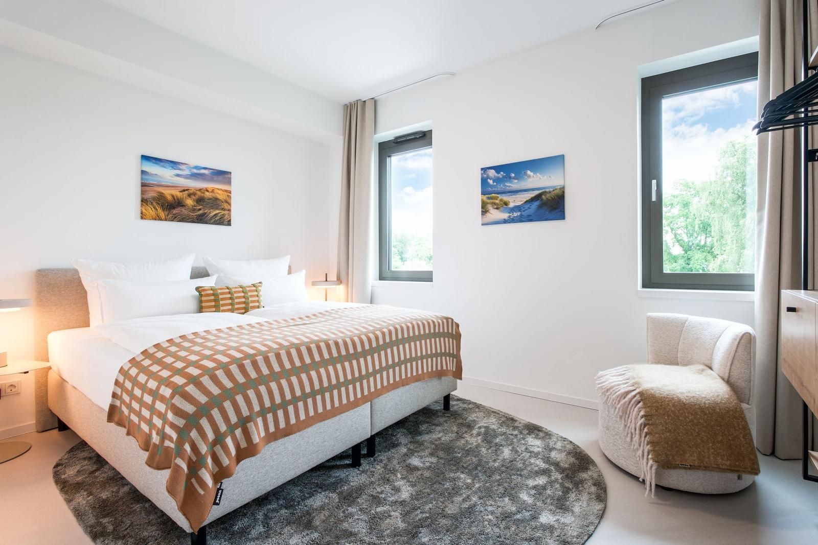Schlafzimmer mit Bett, Stuhl und Fenstern. Wandbilder zeigen Strandlandschaften.