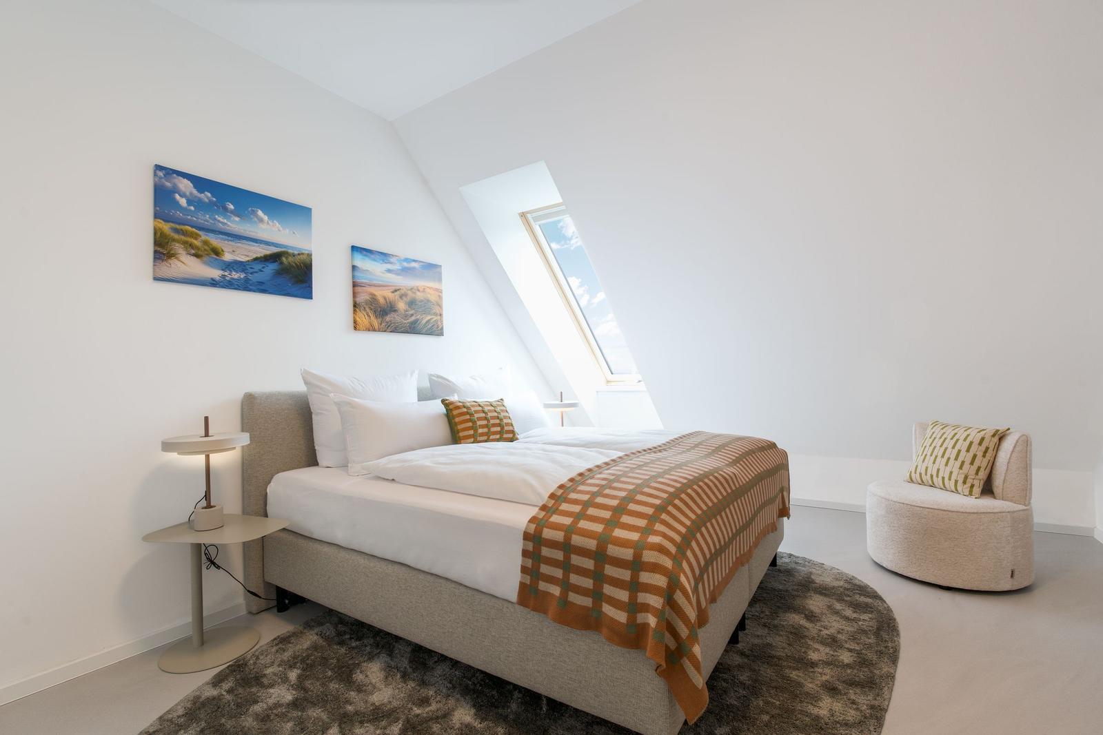Schlafzimmer mit Dachschräge, Bett, Sessel und Wandbildern.