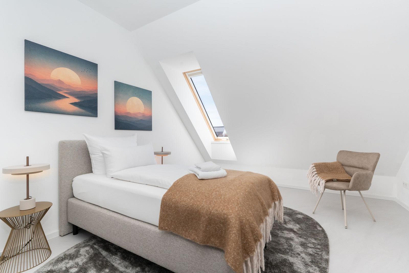 Schlafzimmer mit Dachschrägen, Bett, Sessel und Fenster. Zwei Bilder an der Wand.