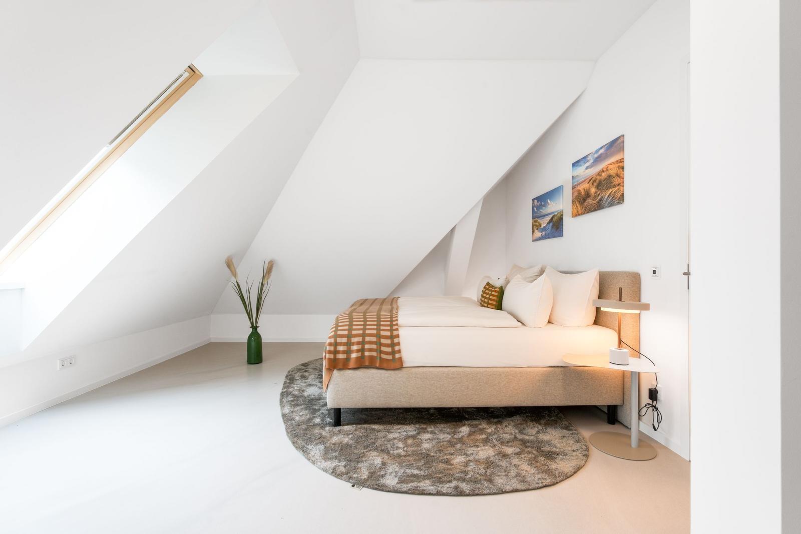 Schlafzimmer mit Dachschrägen, Bett, Teppich und Wandbildern.