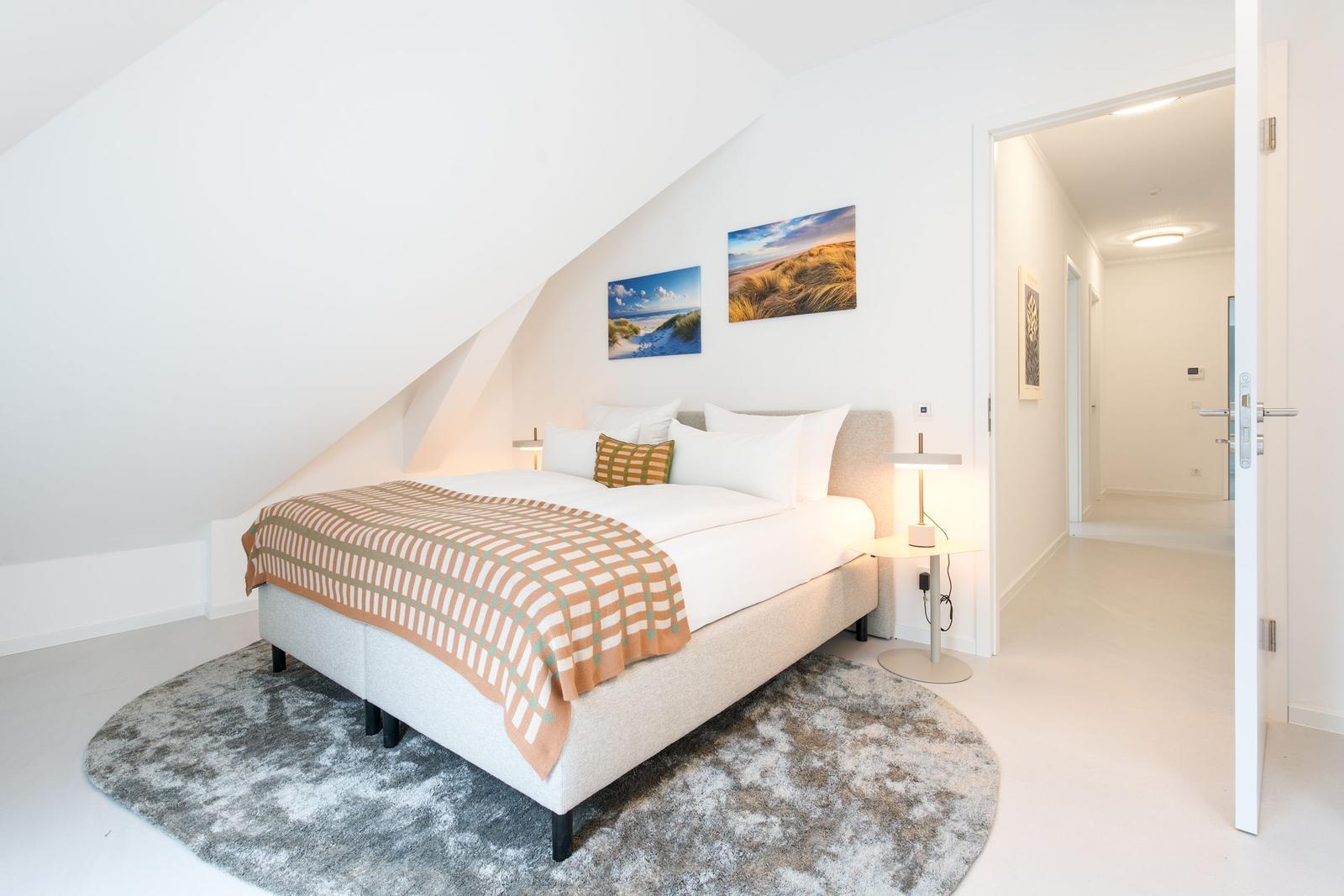 Schlafzimmer mit Dachschräge, Bett, Teppich und Wandbildern. Tür führt in Flur.