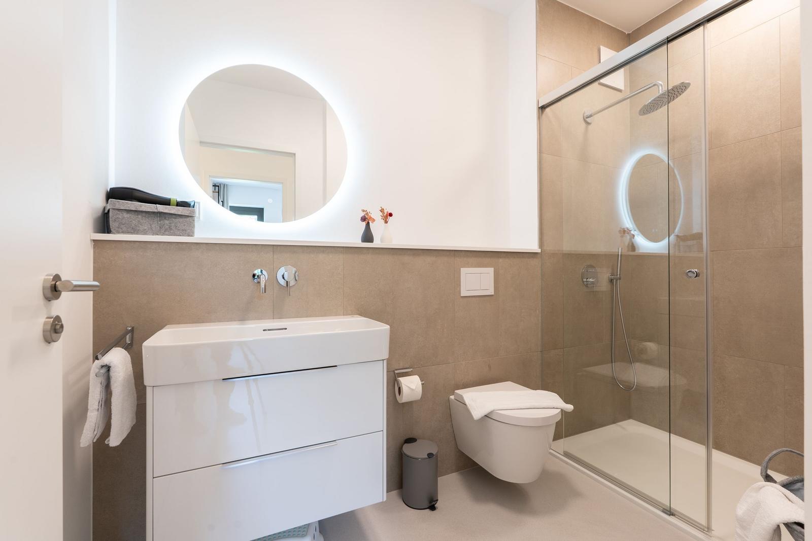 Badezimmer mit Dusche, Waschbecken und Toilette. Rundes Spiegellicht und beige Fliesen.