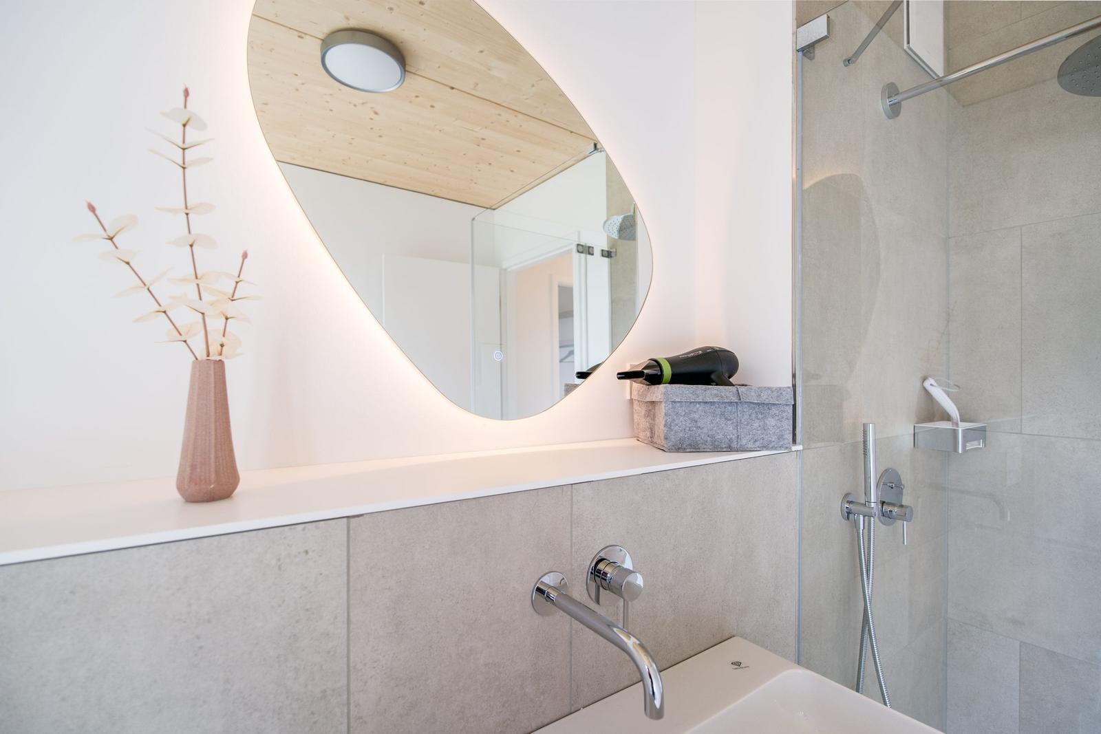 Badezimmer mit Waschbecken, Dusche und beleuchtetem Spiegel.