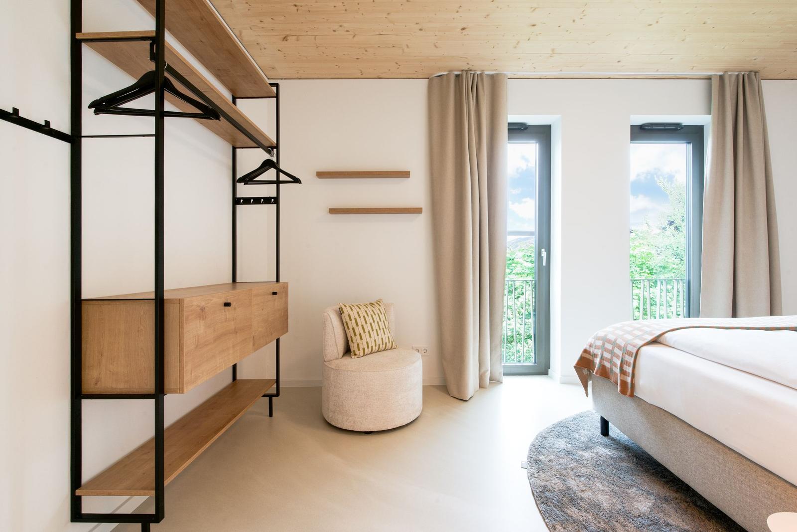 Schlafzimmer mit Bett, Kleiderschrank und Sitzgelegenheit. Fenster mit Blick auf Grün.