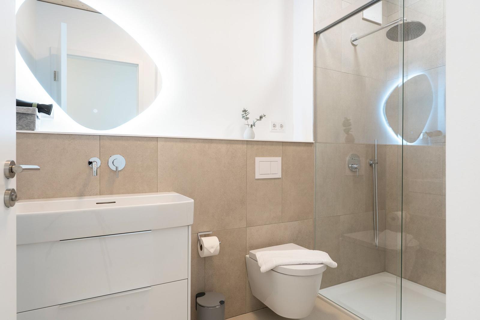 Badezimmer mit Dusche, Waschbecken und Toilette. Moderne Einrichtung mit beleuchtetem Spiegel.
