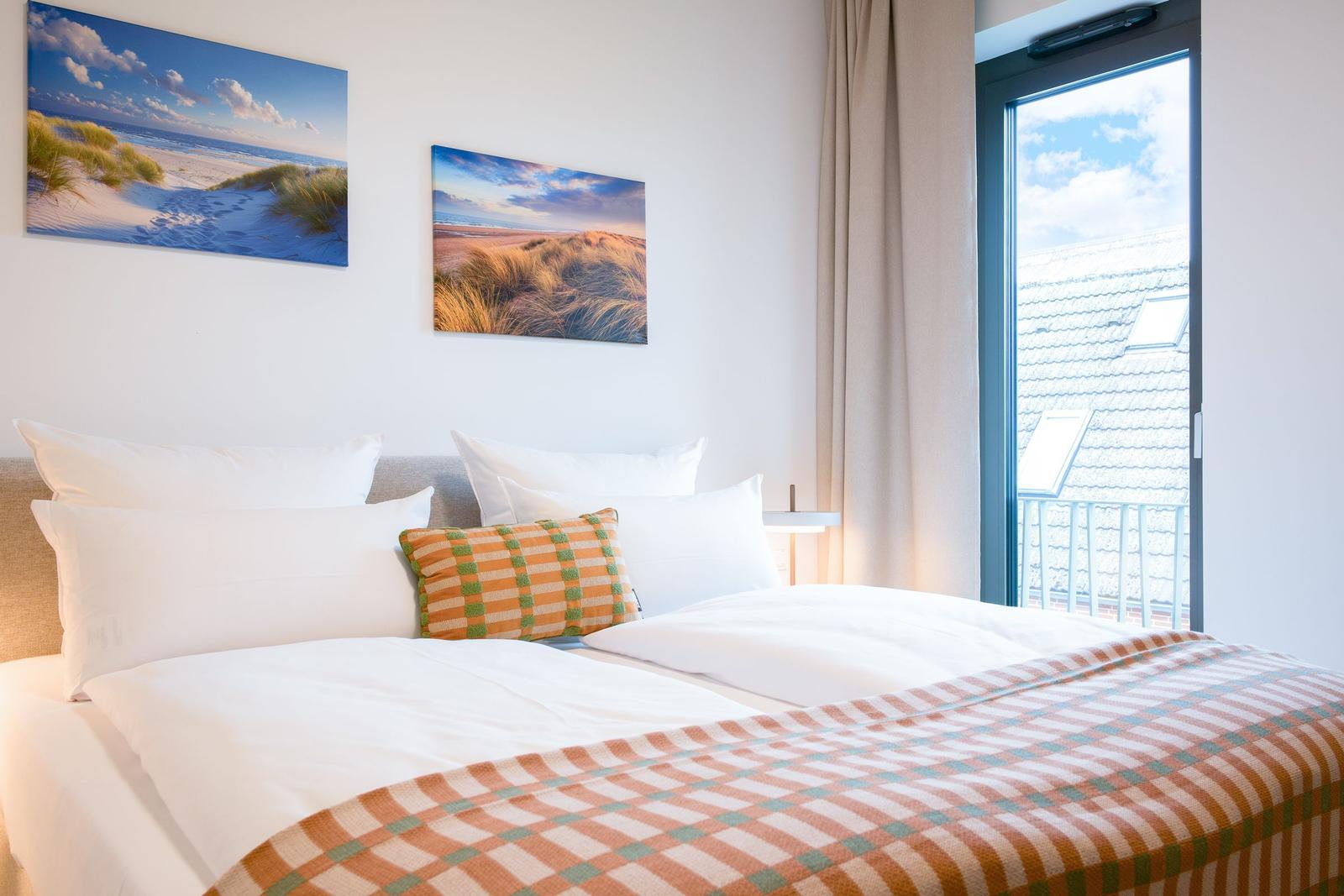 Doppelbett mit Kissen und Decke, Wandbilder und Fenster mit Balkonansicht.