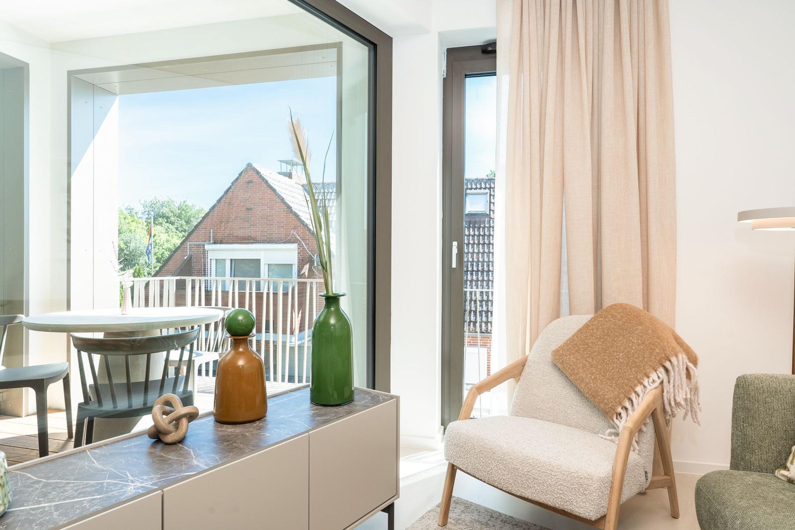 Ein Raum mit Sessel, Tisch und Blick auf Balkon. Vorhänge und Vasen sind sichtbar.