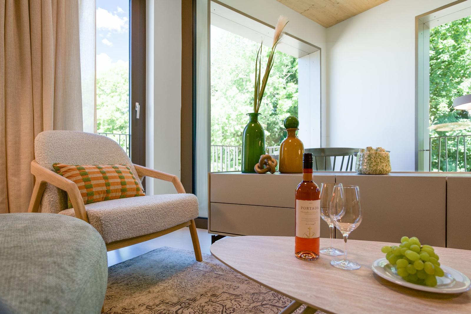 Ein Raum mit Sessel, Tisch und Blick auf Balkon und Grün. Auf Tisch steht Wein und Trauben.