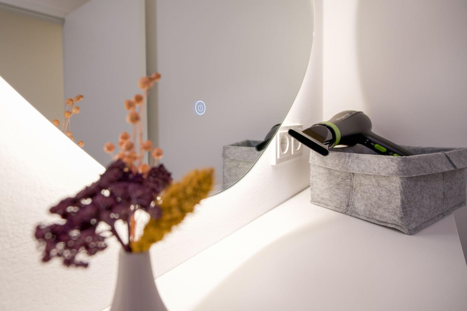 Spiegel mit Touch-Display, Haartrockner und Trockenblumen auf einem weißen Schrank.