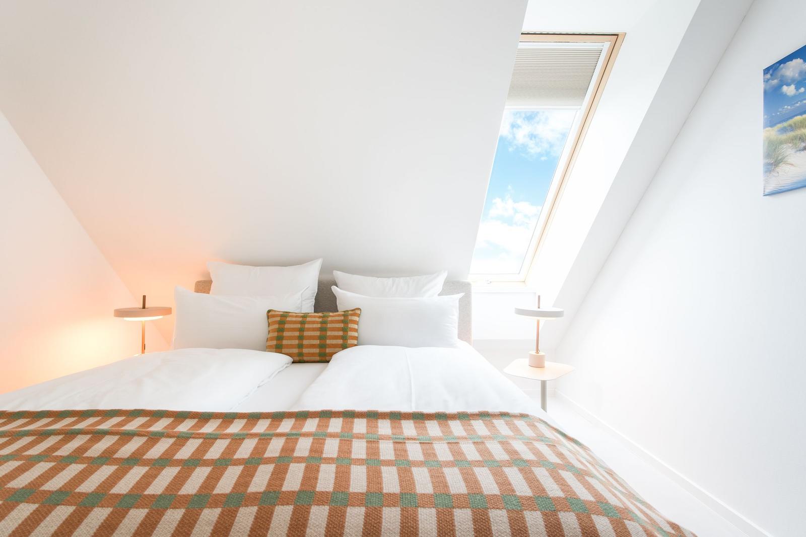 Schlafzimmer mit Dachfenster, Bett mit Kissen und Decke, zwei Nachttische mit Lampen.