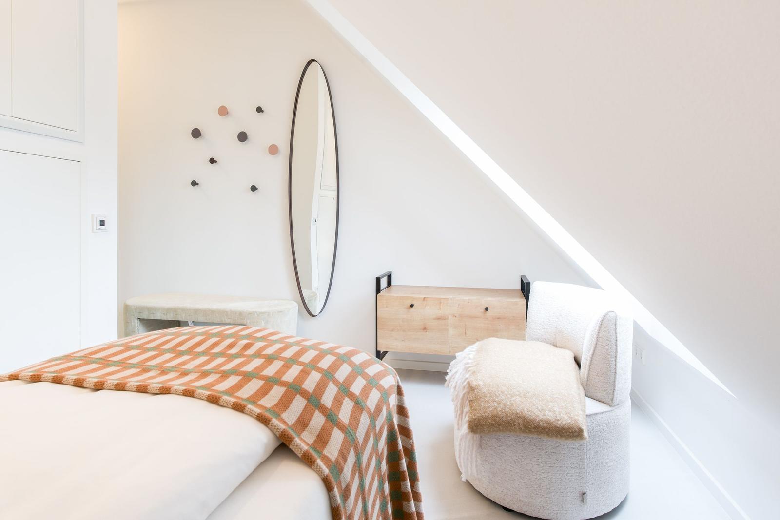 Schlafzimmer mit Bett, Sessel und Spiegel an schräger Wand.