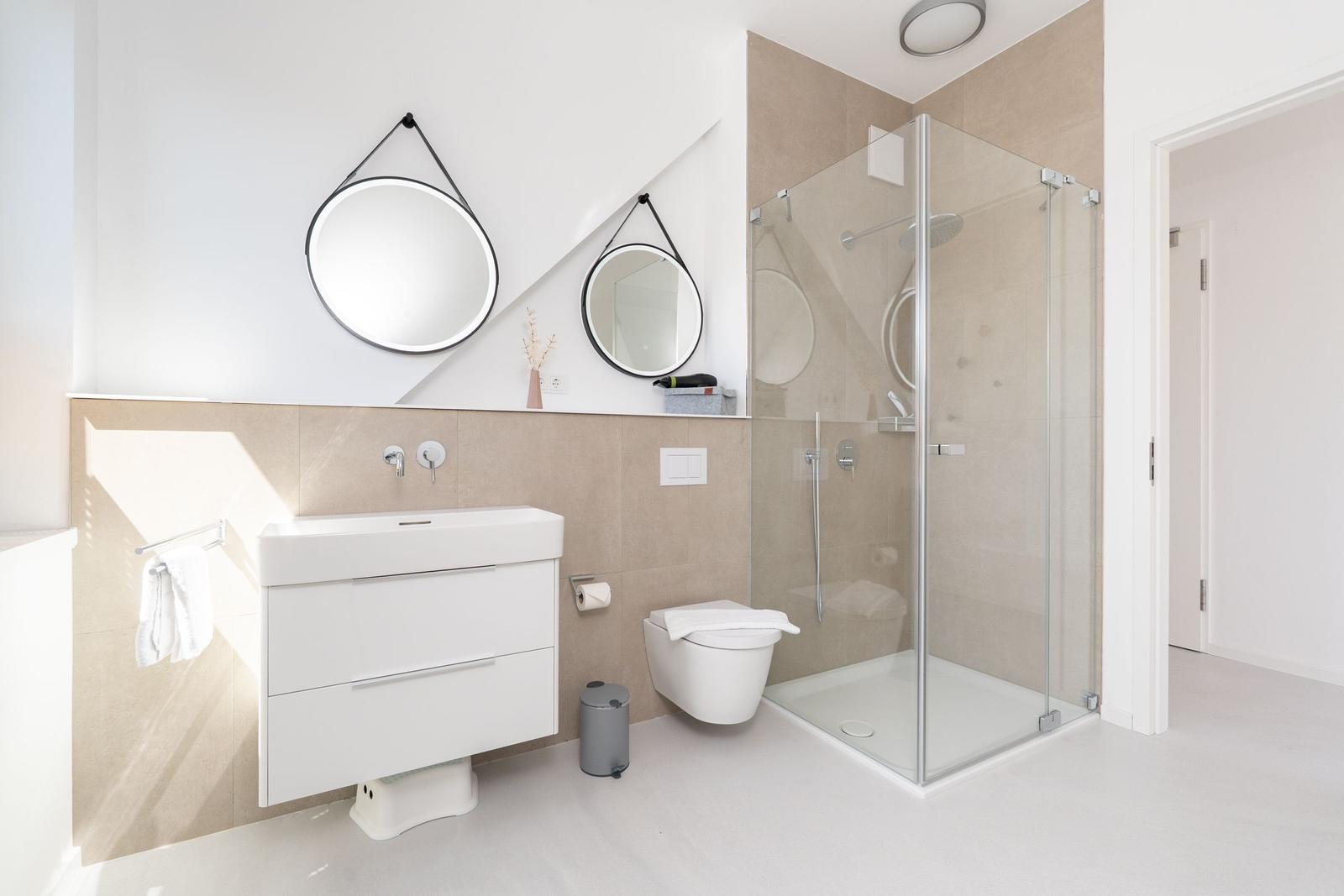 Badezimmer mit Dusche, Waschbecken und Toilette. Zwei runde Spiegel an der Wand.