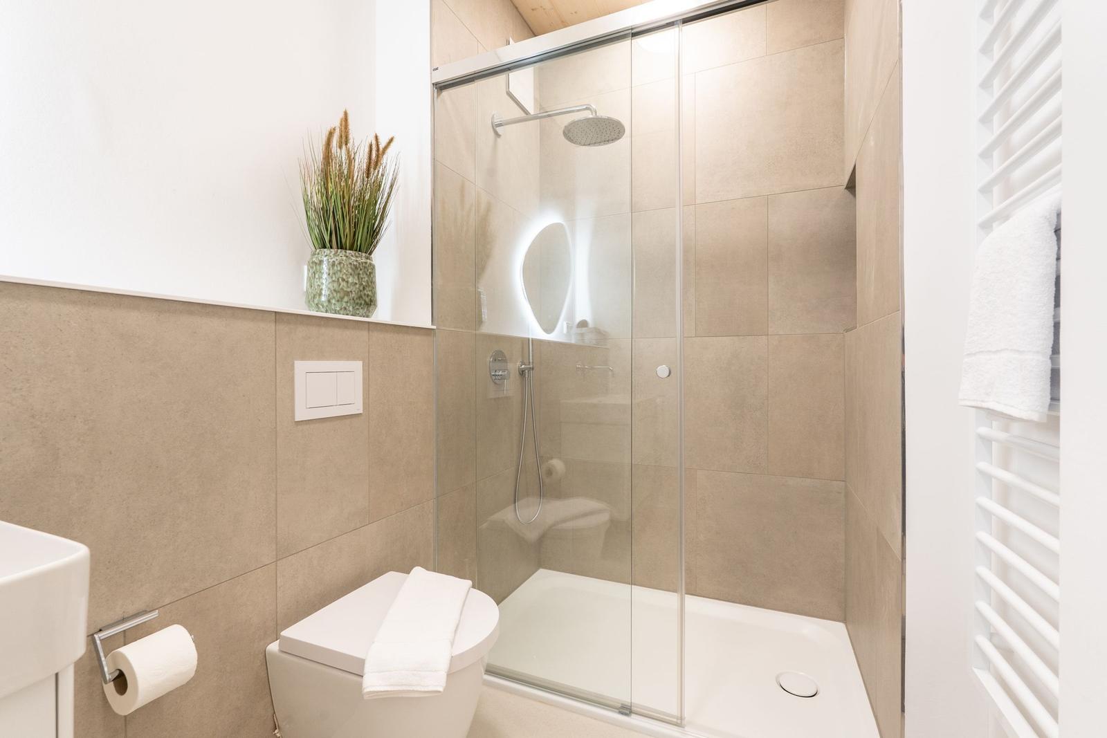 Badezimmer mit Dusche, Toilette und Waschbecken. Weiße Fliesen und Pflanze.