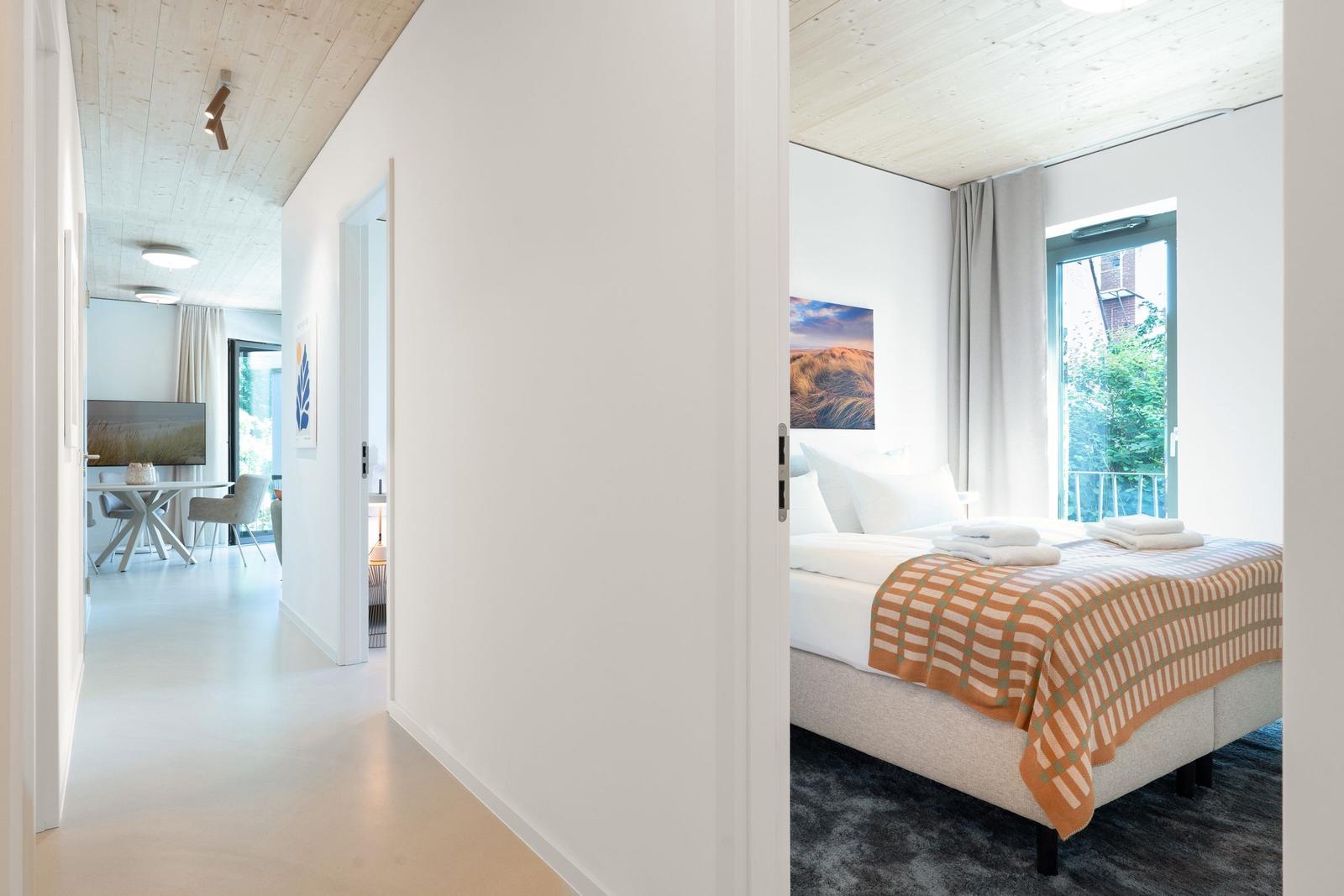 Schlafzimmer mit Bett, Fenster und Decke aus Holz. Durchgang zu Wohnbereich mit Tisch und Stühlen.