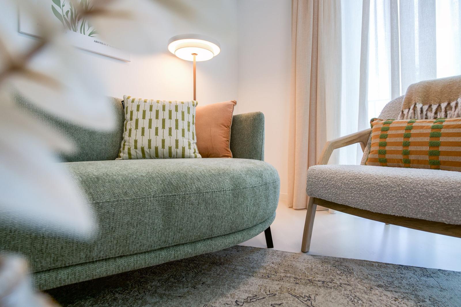 Ein Wohnbereich mit Sofa und Sessel, Bezug in Grün und Beige, Kissen mit Muster, Wandlampe und Fenster mit Vorhängen.